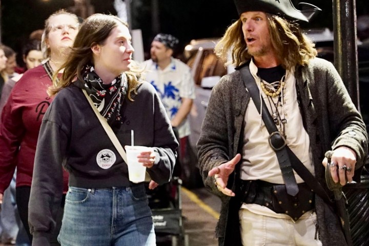 People walking on a street, one dressed as a pirate, talking and holding drinks.